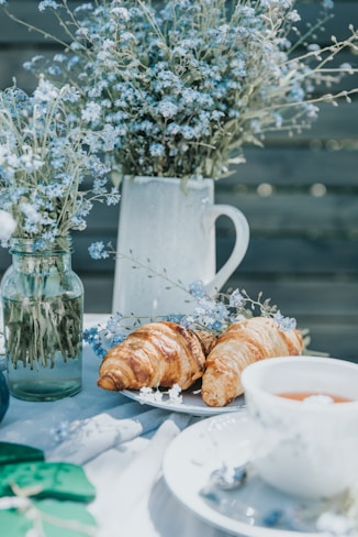 A peaceful outdoor setting featuring a table adorned with delicate blue flowers in glass jars and a ceramic pitcher. Fresh croissants rest on a white plate beside a cup of tea, creating a serene breakfast or brunch atmosphere.