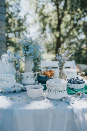 An elegantly set outdoor table features a variety of fine china decorated with floral patterns. Several white ceramic dishes, cups, and a teapot are arranged around the table, accompanied by glass vases filled with delicate blue flowers. Bowls of fresh blueberries and a few pastries add to the scene, which is bathed in soft, natural light. The background reveals a blurred view of green foliage.