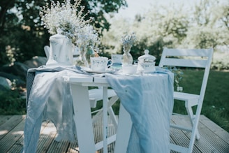 A quaint outdoor table setting with a rustic charm. The white wooden table and chairs are set on a wooden deck, surrounded by lush greenery. The table is elegantly draped with a soft blue linen cloth and adorned with delicate white and blue crockery. A teapot, cups, and a sugar bowl with blue floral patterns complement the look. Fresh blue and white flowers in a decorative vase add a touch of nature and romance.