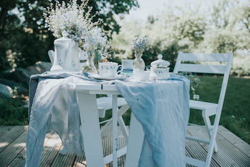 A quaint outdoor table setting with a rustic charm. The white wooden table and chairs are set on a wooden deck, surrounded by lush greenery. The table is elegantly draped with a soft blue linen cloth and adorned with delicate white and blue crockery. A teapot, cups, and a sugar bowl with blue floral patterns complement the look. Fresh blue and white flowers in a decorative vase add a touch of nature and romance.