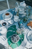 A beautifully arranged tea table with white ceramics including teapots, cups, and a creamer set against a light tablecloth. The table is adorned with sprigs of delicate blue flowers in glass jars. A small bowl of blueberries sits on a large green leaf-shaped placemat, and next to it, a plate holds a couple of croissants.