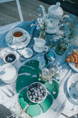 A beautifully arranged tea table with white ceramics including teapots, cups, and a creamer set against a light tablecloth. The table is adorned with sprigs of delicate blue flowers in glass jars. A small bowl of blueberries sits on a large green leaf-shaped placemat, and next to it, a plate holds a couple of croissants.