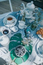 A beautifully arranged tea table with white ceramics including teapots, cups, and a creamer set against a light tablecloth. The table is adorned with sprigs of delicate blue flowers in glass jars. A small bowl of blueberries sits on a large green leaf-shaped placemat, and next to it, a plate holds a couple of croissants.