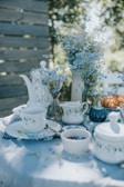 A serene outdoor tea setting featuring porcelain teacups and a teapot adorned with blue floral patterns. The table is decorated with delicate blue wildflowers in glass vases, and various items like a bowl of jam, a covered dish, and croissants are placed on a white tablecloth. Soft, dappled sunlight filters through the trees, creating a peaceful and inviting atmosphere.