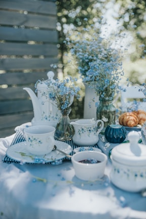 A serene outdoor tea setting featuring porcelain teacups and a teapot adorned with blue floral patterns. The table is decorated with delicate blue wildflowers in glass vases, and various items like a bowl of jam, a covered dish, and croissants are placed on a white tablecloth. Soft, dappled sunlight filters through the trees, creating a peaceful and inviting atmosphere.