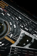 An aerial view of a multi-lane road intersection with several cars in various lanes, some waiting at pedestrian crossings marked by yellow stripes. The scene includes curved and straight road sections, traffic signs, and green patches of grass with trees.
