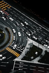 An aerial view of a multi-lane road intersection with several cars in various lanes, some waiting at pedestrian crossings marked by yellow stripes. The scene includes curved and straight road sections, traffic signs, and green patches of grass with trees.