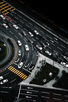 An aerial view of a multi-lane road intersection with several cars in various lanes, some waiting at pedestrian crossings marked by yellow stripes. The scene includes curved and straight road sections, traffic signs, and green patches of grass with trees.