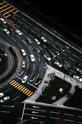 An aerial view of a multi-lane road intersection with several cars in various lanes, some waiting at pedestrian crossings marked by yellow stripes. The scene includes curved and straight road sections, traffic signs, and green patches of grass with trees.