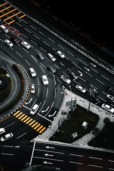 An aerial view of a multi-lane road intersection with several cars in various lanes, some waiting at pedestrian crossings marked by yellow stripes. The scene includes curved and straight road sections, traffic signs, and green patches of grass with trees.