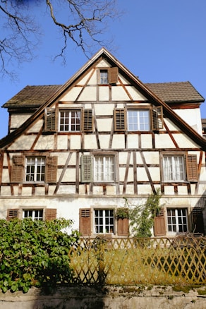 A charming timber-framed house with a steeply pitched roof, featuring multiple windows with wooden shutters. The building has a traditional design with ivy growing on the fa&ccedil;ade. In the foreground, there is a wooden trellis fence surrounded by green foliage. The sky is clear and blue, with a few bare tree branches visible.
