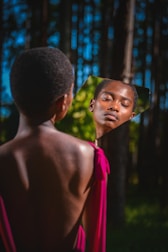 A person stands in a forested area, looking into a fragmented mirror that reflects their face. Sunlight filters through the trees, casting a warm glow. The person wears a vibrant pink garment with an exposed back, and the surroundings are lush and green.