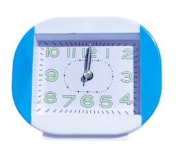 A square-shaped clock with a blue frame and white face. The clock displays numbers in a stylized font, highlighted with green. The hands of the clock are silver, pointing towards 10 and 12.