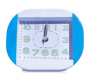 A square-shaped clock with a blue frame and white face. The clock displays numbers in a stylized font, highlighted with green. The hands of the clock are silver, pointing towards 10 and 12.
