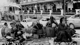 A group of women wearing traditional clothing are seated on the ground with various handmade items and baskets around them. The scene takes place on a busy street with cars parked nearby, and there is a backdrop of shops with decorative lights. The mood feels lively but also contemplative, as the women appear to be engaged in conversation or thought.
