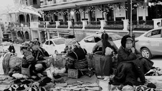 A group of women wearing traditional clothing are seated on the ground with various handmade items and baskets around them. The scene takes place on a busy street with cars parked nearby, and there is a backdrop of shops with decorative lights. The mood feels lively but also contemplative, as the women appear to be engaged in conversation or thought.