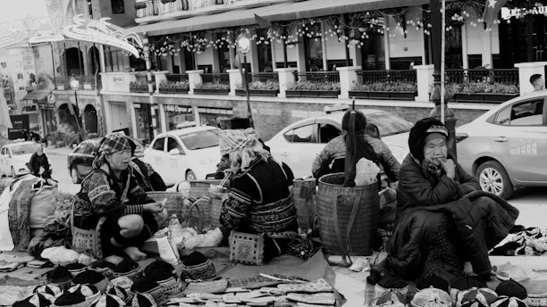 A group of women wearing traditional clothing are seated on the ground with various handmade items and baskets around them. The scene takes place on a busy street with cars parked nearby, and there is a backdrop of shops with decorative lights. The mood feels lively but also contemplative, as the women appear to be engaged in conversation or thought.