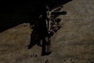 A crucifix hangs on a textured, aged wall with shadow patterns. The image has a somber atmosphere with minimal light, creating a dramatic contrast. Latin numerals and some indecipherable text are visible on the wall beside the crucifix.