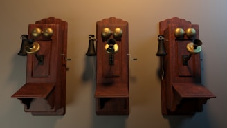 Three vintage wall-mounted telephones with wooden bases and brass components, including a speaker, receiver, and crank handle, are arranged side by side against a plain backdrop. The telephones appear to be from the early 20th century, with intricate craftsmanship and a warm patina on the wood and brass.