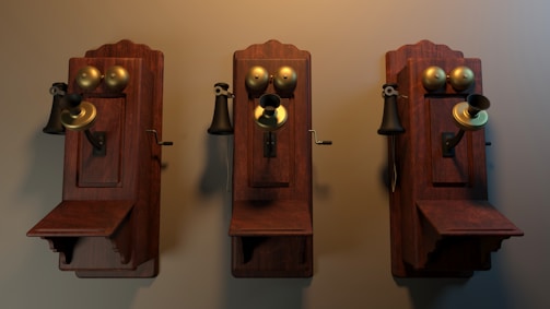 Three vintage wall-mounted telephones with wooden bases and brass components, including a speaker, receiver, and crank handle, are arranged side by side against a plain backdrop. The telephones appear to be from the early 20th century, with intricate craftsmanship and a warm patina on the wood and brass.