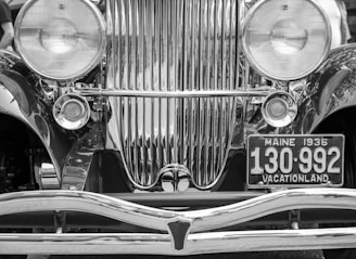A close-up of the front of a vintage car featuring prominent chrome detailing. The vehicle has large round headlights on either side of a vertical grille with shiny bars. A license plate reads 'Maine 1936 130-992' with the words 'Vacationland' displayed below. The image is in black and white, highlighting the reflective surfaces and intricate design.