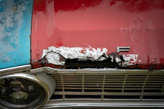 A close-up view of the front section of an old car showing significant rust damage. The hood is primarily painted red with a worn spot revealing metal corrosion beneath. Adjacent areas show peeling paint and discoloration, with a distinctive emblem featuring the letter 'F' prominently visible. The headlight on the left is rounded and slightly tarnished, surrounded by a metal frame.