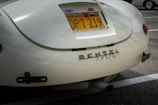 A close-up of the rear section of a vintage car with a California license plate. The car's paint appears to be a cream color and the back features a chrome emblem spelling 'RENZEL 1300'. The car is parked in a lot, with another car partially visible in the background.