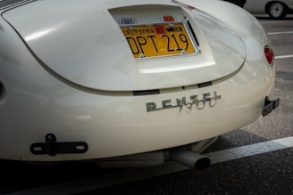 A close-up of the rear section of a vintage car with a California license plate. The car's paint appears to be a cream color and the back features a chrome emblem spelling 'RENZEL 1300'. The car is parked in a lot, with another car partially visible in the background.