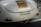 A close-up of the rear section of a vintage car with a California license plate. The car's paint appears to be a cream color and the back features a chrome emblem spelling 'RENZEL 1300'. The car is parked in a lot, with another car partially visible in the background.