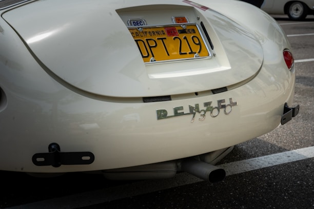 A close-up of the rear section of a vintage car with a California license plate. The car's paint appears to be a cream color and the back features a chrome emblem spelling 'RENZEL 1300'. The car is parked in a lot, with another car partially visible in the background.