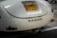 A close-up of the rear section of a vintage car with a California license plate. The car's paint appears to be a cream color and the back features a chrome emblem spelling 'RENZEL 1300'. The car is parked in a lot, with another car partially visible in the background.