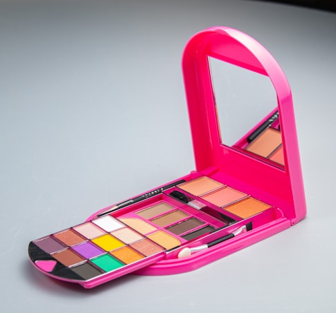 A brightly colored pink makeup palette with a mirror. The palette includes multiple compartments containing a variety of eyeshadows in shades like purple, orange, yellow, and green. There are also makeup brushes included in the set.