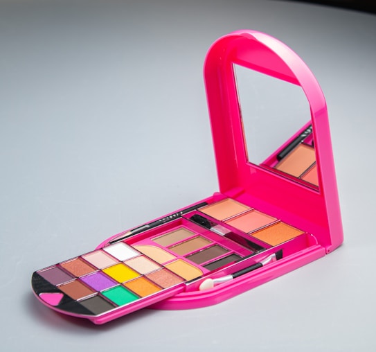 A brightly colored pink makeup palette with a mirror. The palette includes multiple compartments containing a variety of eyeshadows in shades like purple, orange, yellow, and green. There are also makeup brushes included in the set.