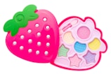 A pink, strawberry-shaped makeup kit with a green leafy top, featuring compartments with pastel-colored eyeshadows and shapes like stars and circles. The design is playful and colorful, aimed at children.