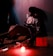 A person is sitting on the floor leaning against a wall. The red lighting casts an intense hue over the scene, creating a dramatic atmosphere. The figure appears to be holding a large gun and is wearing a white shirt with red stains. Near the person, a red light bulb is glowing on the floor alongside various scattered items, including a book with the title 'How to Get Away with Murder' and a knife. The setting feels tense and ominous.