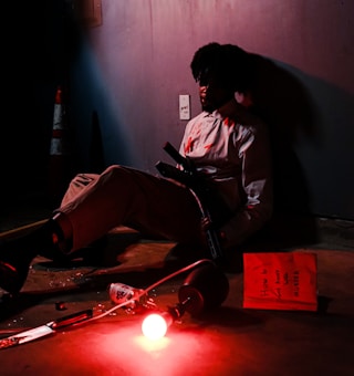 A person is sitting on the floor leaning against a wall. The red lighting casts an intense hue over the scene, creating a dramatic atmosphere. The figure appears to be holding a large gun and is wearing a white shirt with red stains. Near the person, a red light bulb is glowing on the floor alongside various scattered items, including a book with the title 'How to Get Away with Murder' and a knife. The setting feels tense and ominous.