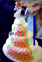 A multi-tiered cake covered with pink rose-shaped decorations and green leaves. The cake has a neat arrangement of small, round embellishments and is topped with a bride and groom figurine holding a bouquet of flowers. The design is intricate and elegant, suitable for a wedding.