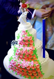 A multi-tiered cake covered with pink rose-shaped decorations and green leaves. The cake has a neat arrangement of small, round embellishments and is topped with a bride and groom figurine holding a bouquet of flowers. The design is intricate and elegant, suitable for a wedding.