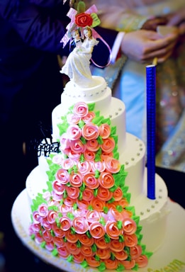 A multi-tiered cake covered with pink rose-shaped decorations and green leaves. The cake has a neat arrangement of small, round embellishments and is topped with a bride and groom figurine holding a bouquet of flowers. The design is intricate and elegant, suitable for a wedding.