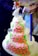 A multi-tiered cake covered with pink rose-shaped decorations and green leaves. The cake has a neat arrangement of small, round embellishments and is topped with a bride and groom figurine holding a bouquet of flowers. The design is intricate and elegant, suitable for a wedding.