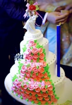 A multi-tiered cake covered with pink rose-shaped decorations and green leaves. The cake has a neat arrangement of small, round embellishments and is topped with a bride and groom figurine holding a bouquet of flowers. The design is intricate and elegant, suitable for a wedding.