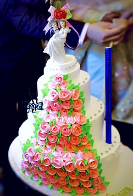 A multi-tiered cake covered with pink rose-shaped decorations and green leaves. The cake has a neat arrangement of small, round embellishments and is topped with a bride and groom figurine holding a bouquet of flowers. The design is intricate and elegant, suitable for a wedding.
