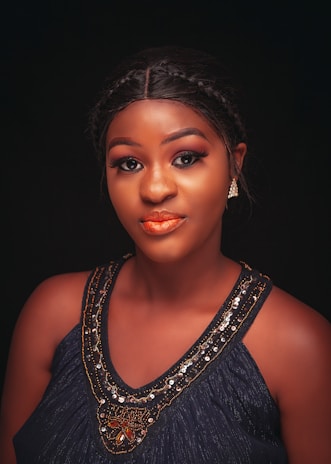 A woman with braided hair and elegant makeup poses against a dark background. She wears a richly embellished dark top with ornate beadwork and looks confidently at the camera. Her lips have a vibrant orange shade, and she has earrings adding to her stylish appearance.