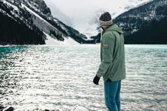 A model wearing a deep green sweatshirt standing beside a serene mountain lake at dawn.