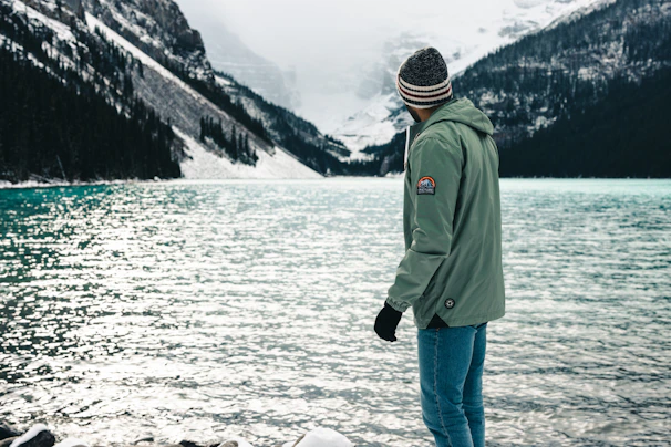 A model wearing a deep green sweatshirt standing beside a serene mountain lake at dawn.