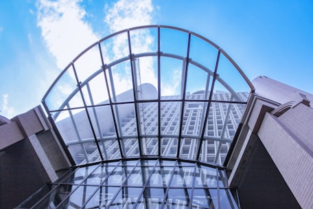 A modern architectural structure viewed from below, featuring an intricate grid of glass and steel with a prominent arch over a building facade. The structure is set against a bright blue sky with scattered clouds.