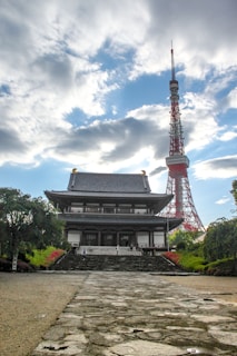 A traditional Japanese temple stands prominently in the foreground, featuring detailed wooden architecture with tiled roofs. Behind the temple, a tall red and white communications tower rises into a partly cloudy sky. The setting is serene with well-maintained gardens and pathways leading to the temple entrance.