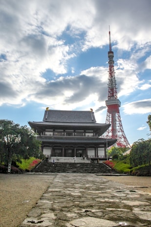 A traditional Japanese temple stands prominently in the foreground, featuring detailed wooden architecture with tiled roofs. Behind the temple, a tall red and white communications tower rises into a partly cloudy sky. The setting is serene with well-maintained gardens and pathways leading to the temple entrance.