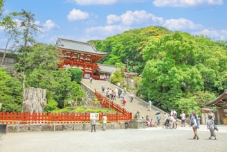 A traditional Japanese temple with vibrant red architecture sits atop a wide staircase, surrounded by lush green trees and a bright blue sky with scattered clouds. Numerous visitors, including families and couples, wander around, some ascending the stairs, others taking photos. The foreground features a red wooden fence and a large stone lantern.