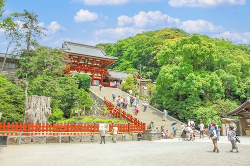 A traditional Japanese temple with vibrant red architecture sits atop a wide staircase, surrounded by lush green trees and a bright blue sky with scattered clouds. Numerous visitors, including families and couples, wander around, some ascending the stairs, others taking photos. The foreground features a red wooden fence and a large stone lantern.